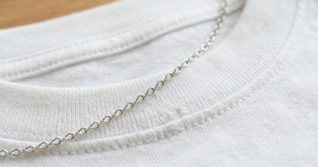 白いコットンのTシャツの首元にシルバーの細いチェーンが重なり、リブ部分にわずかな毛羽立ちや傷みが見える接写。