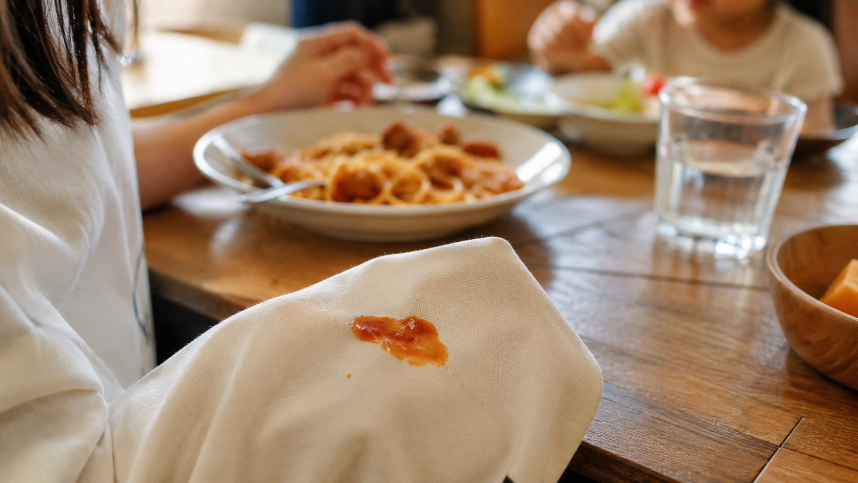 家族の食卓で起きる「あーっ!」の瞬間。ミートソースやラーメンのスープが白い布(ナプキンや服の袖)にこぼれた直後のクローズアップ。子育てや家族のいる暮らしで誰もが経験する食べこぼしのあるあるシーン。北九州市小倉南区のファミリークリーニング「諦めないでシリーズ」第8弾の導入画像。